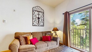 A living room with a sofa, a lamp, and a view of trees through a window. - Sweet Retreat - Ski Valley Condo, Gym, Game Room (Taos Ski Valley)