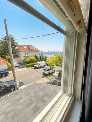Blick aus einem Fenster auf eine Straße mit parkenden Autos, ein Gebäude mit rotem Dach und eine ferne Stadtlandschaft.