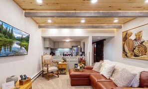 A living room with a brown leather sofa, a wooden chair, a television, and a kitchen in the background.
