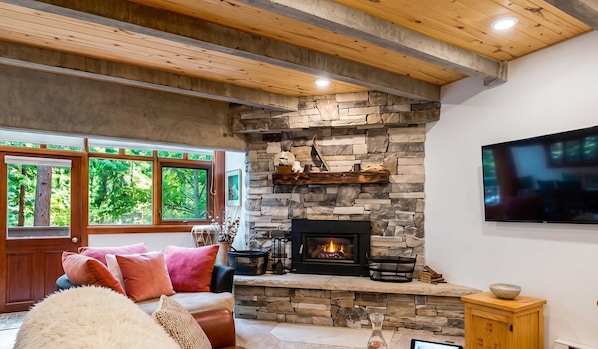 A living room with a stone fireplace, large windows, a flat-screen TV, and a cozy seating area.
