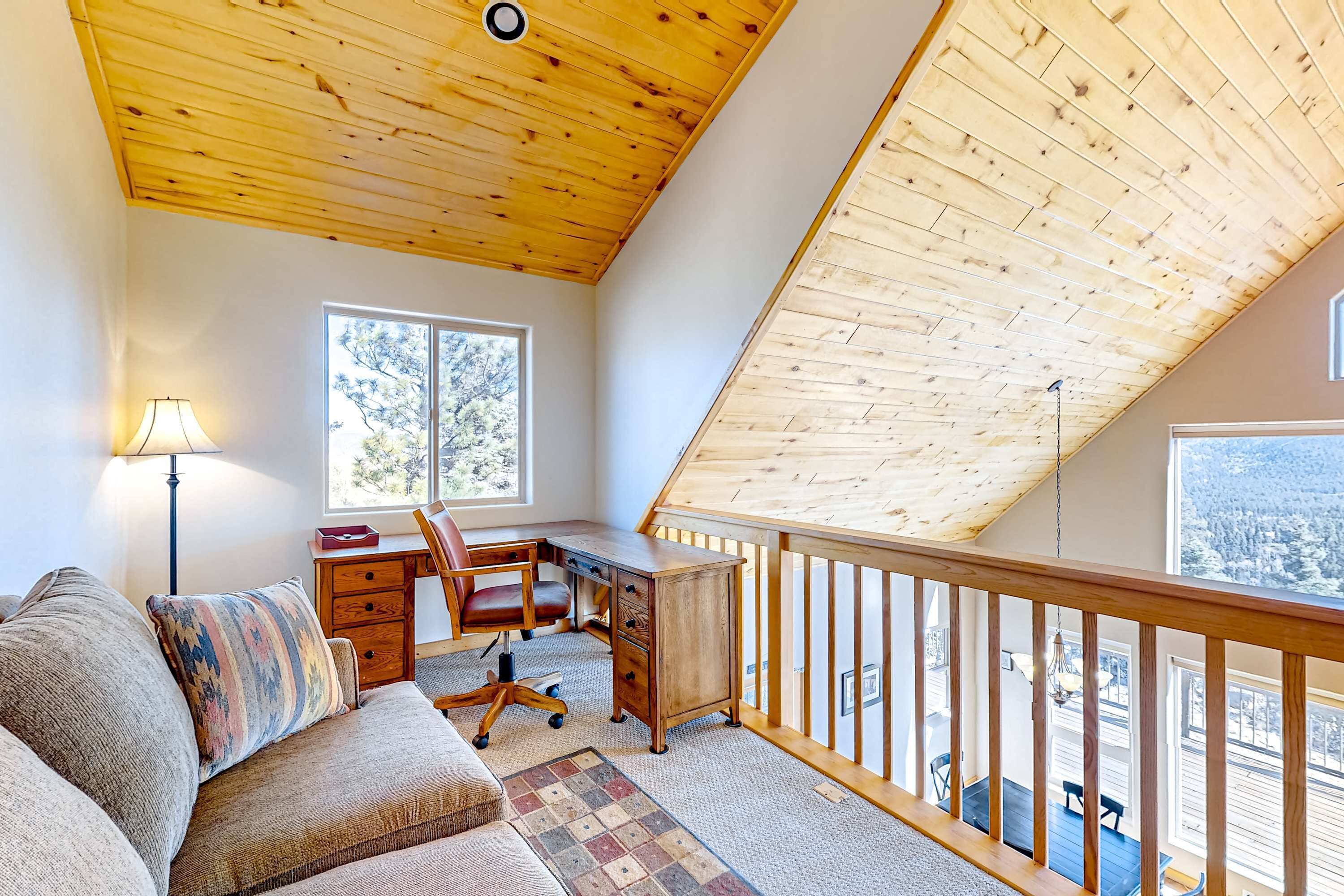 A loft-style room with a wooden ceiling, a desk with a chair, a sofa, and a view of the outdoors through large windows.