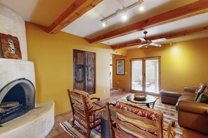 A living room with a fireplace, wooden ceiling beams, a ceiling fan, and a large window with blinds. - Susan's Hideaway - Santa Fe Charm & Walk to Plaza (Santa Fe)