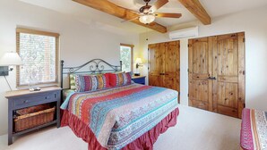 Une chambre à coucher avec un lit, des poutres en bois au plafond, un ventilateur de plafond, une table de chevet, une lampe, un panier en osier et des armoires en bois.