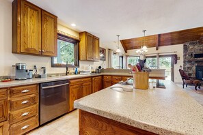 A kitchen with wooden cabinets, a dishwasher, granite countertops, and a stone fireplace. - Pine Valley Lodge - Views, Deck, Firepit & Sauna (Angel Fire)