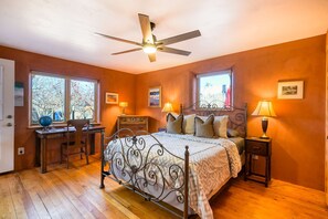 Ein Schlafzimmer mit Bett, Schreibtisch, Stuhl, zwei Fenstern, Deckenventilator und Nachttischlampen.