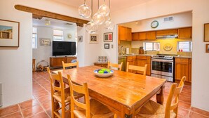 A dining area with a wooden table, chairs, and a bowl of fruit. There is a kitchen with wooden cabinets, a stove, and a refrigerator. - Santa Fe Serenity - Walk to the Railyard, Dog-Friendly Garden Home (Santa Fe)