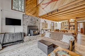 A living room with a stone fireplace, a sofa, a television, and a kitchen in the background. - Timbers Townhouse 1 - Walk to Lifts, Hike & Bike (Angel Fire)