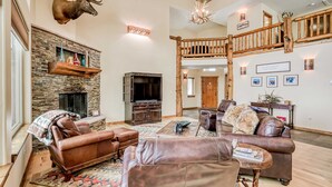 A spacious living room with a stone fireplace, leather sofas, a mounted deer head, and a wooden staircase. - Vista Linda Cabin - Views, Sauna, Deck & Dog Okay (Angel Fire)