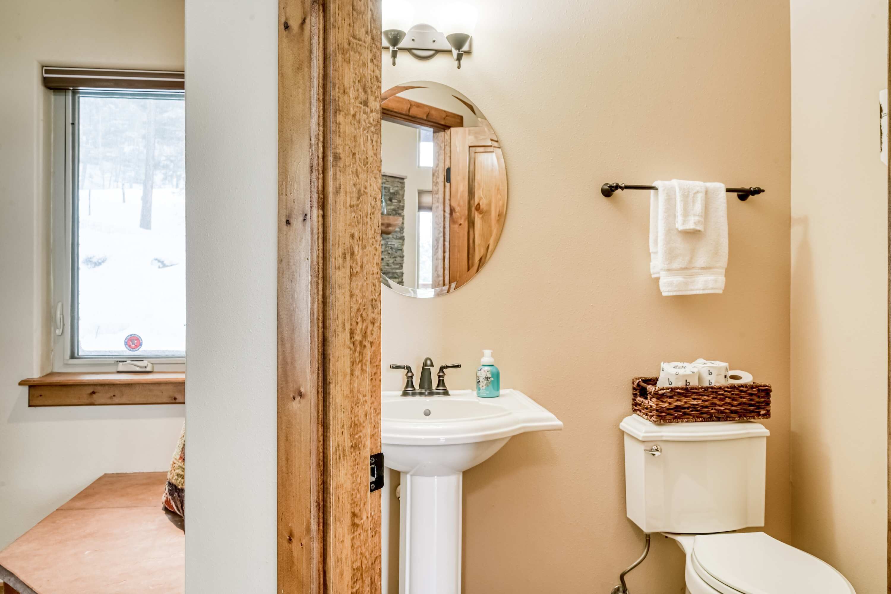 A bathroom with a wooden door, a round mirror, a sink, a toilet, and a window with a view of trees.
