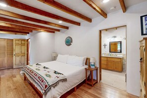 Ein Schlafzimmer mit einem großen Bett, Holzbalken, einem Nachttisch, einer Wanduhr und einem durchgegangenen Tür angedeuteten Badezimmer.