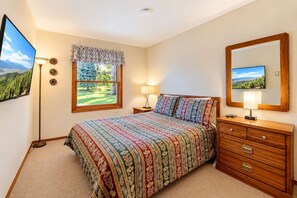 Ein Schlafzimmer mit einem Bett, einer Kommode, einem Nachttisch, einer Lampe, einem Fenster mit Landschaftsblick und einem Spiegel.