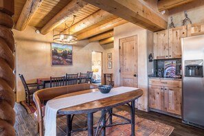 Ein rustikales Interieur mit einem Holzespeisetisch, Stühlen, einer großen Schüssel, einer Chandelüster, Holzschränken und einem Kühlschrank.