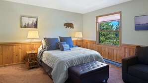 Une chambre à coucher avec un lit, deux tables de chevet avec des lampes, une tête de lit en bois, une fenêtre donnant sur un paysage agréable et une décoration murale représentant un ours.