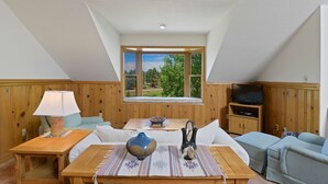 Une pièce avec une table en bois, un canapé, un téléviseur et une fenêtre donnant sur un paysage verdoyant.