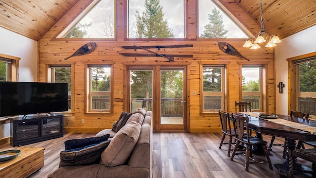 A wooden cabin interior with a TV, sofa, dining table, and large windows. - The Bear Chalet - Hot Tub, Game Room, Mtn Views (Angel Fire)