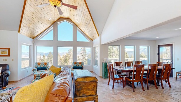 A spacious living area with a high ceiling, large windows, a dining table with chairs, and a leather sofa. - Southwest Heaven - Sweeping Views, Hot Tub & Deck (Angel Fire)