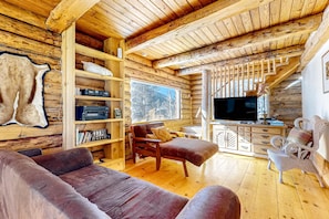 A log cabin interior with a wooden ceiling, a sofa, a chair, a television, and a bookshelf.