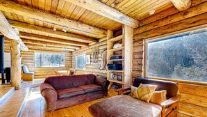A log cabin interior with a wooden ceiling, a brown leather sofa, a wooden bench, and a large window. - Ridgerunner Cabin - Ski Retreat with Hot Tub (Taos Ski Valley)
