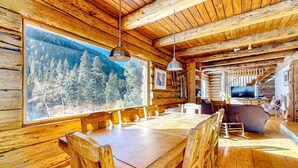 A wooden cabin interior with a large dining table, wooden chairs, a television, and a view of a forest through a large window. - Ridgerunner Cabin - Ski Retreat with Hot Tub (Taos Ski Valley)