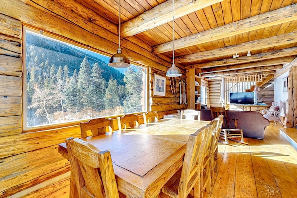 A wooden cabin interior with a large dining table, wooden chairs, a television, and a view of a forest through a large window.