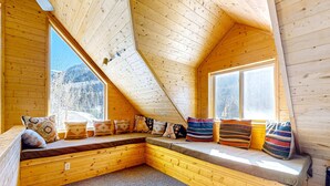A cozy cabin with a sloped ceiling, a built-in bench with cushions, and large windows offering a view of the outdoors. - Ridgerunner Cabin - Ski Retreat with Hot Tub (Taos Ski Valley)
