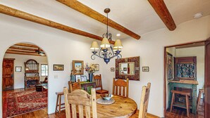 A dining area with a wooden table, chairs, a chandelier, and a red rug. - Casa Raya Taos - Mountain Views, Yard & Near Plaza (Taos)