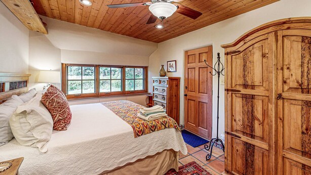 A bedroom with a wooden ceiling, a bed with a patterned bedspread, a large wardrobe, and a window with a view of trees. - Casa Raya Taos - Mountain Views, Yard & Near Plaza (Taos)