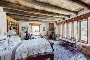 Een rustieke slaapkamer met een houten plafond, een groot bed met een quilt met bloemenprint, een open haard en uitzicht door grote ramen.
