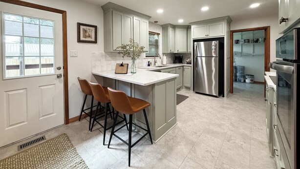 A modern kitchen with a central island, bar stools, a stainless steel refrigerator, and a view into the adjacent living area. - Skyline House - Sunrise Oasis (Searcy)