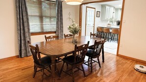 A dining room with a wooden table and chairs, a vase with flowers, and a kitchen visible through an open door. - Skyline House - Sunrise Oasis (Searcy)