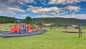 Ein Spielplatz mit Rutschen und Klettergerüsten, umgeben von einer grünen Wiese und einem bewaldeten Hügel im Hintergrund.