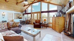 A spacious living room with a high ceiling, wooden beams, a ceiling fan, a fireplace, a dining area, and large windows. - El Camino Real Retreat - Views, Hot Tub & Pets Ok (Angel Fire)