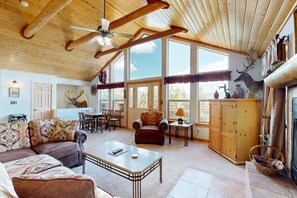 A spacious living room with a high ceiling, wooden beams, a ceiling fan, a fireplace, a dining area, and large windows. - El Camino Real Retreat - Views, Hot Tub & Pets Ok (Angel Fire)