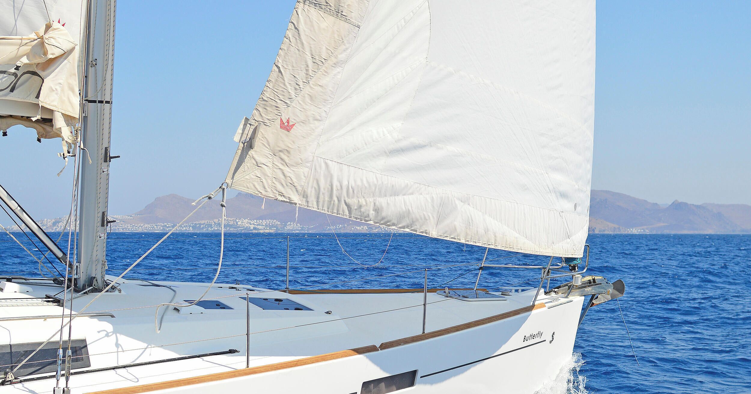 A sailboat with a white hull and a sail, sailing on blue water with a mountainous coastline in the background.