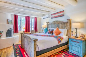 A bedroom with a wooden bed, a red and black patterned rug, a wooden nightstand, a white bedside table, and a window with red curtains. - Casa De Muneco - Adobe Charm, Steps from Canyon Rd (Santa Fe)