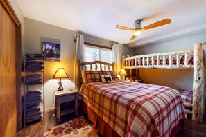Um quarto com cama, mesa de cabeceira, luminária, ventoinha no teto, janela com cortinas, prateleira na parede e piso de madeira.