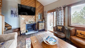 A cozy living room with a fireplace, leather sofas, a coffee table with magazines, a TV mounted on a wooden wall, and a window with curtains. - The Lift C2 - Walk to Lifts & Town, Mountain Views (Angel Fire)