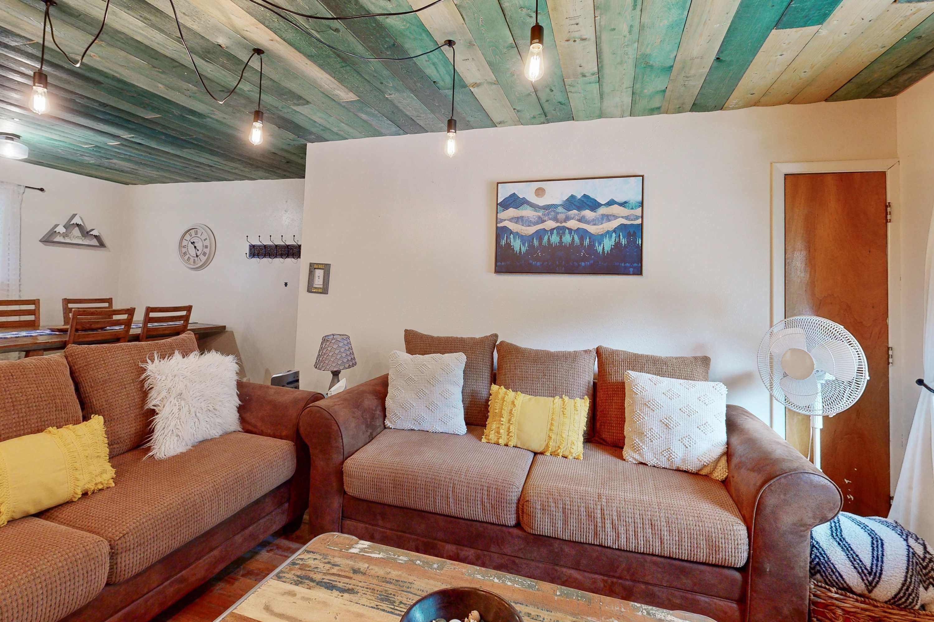 Sala de estar com dois sofás marrons, teto de madeira, relógio de parede, um quadro e um ventilador.