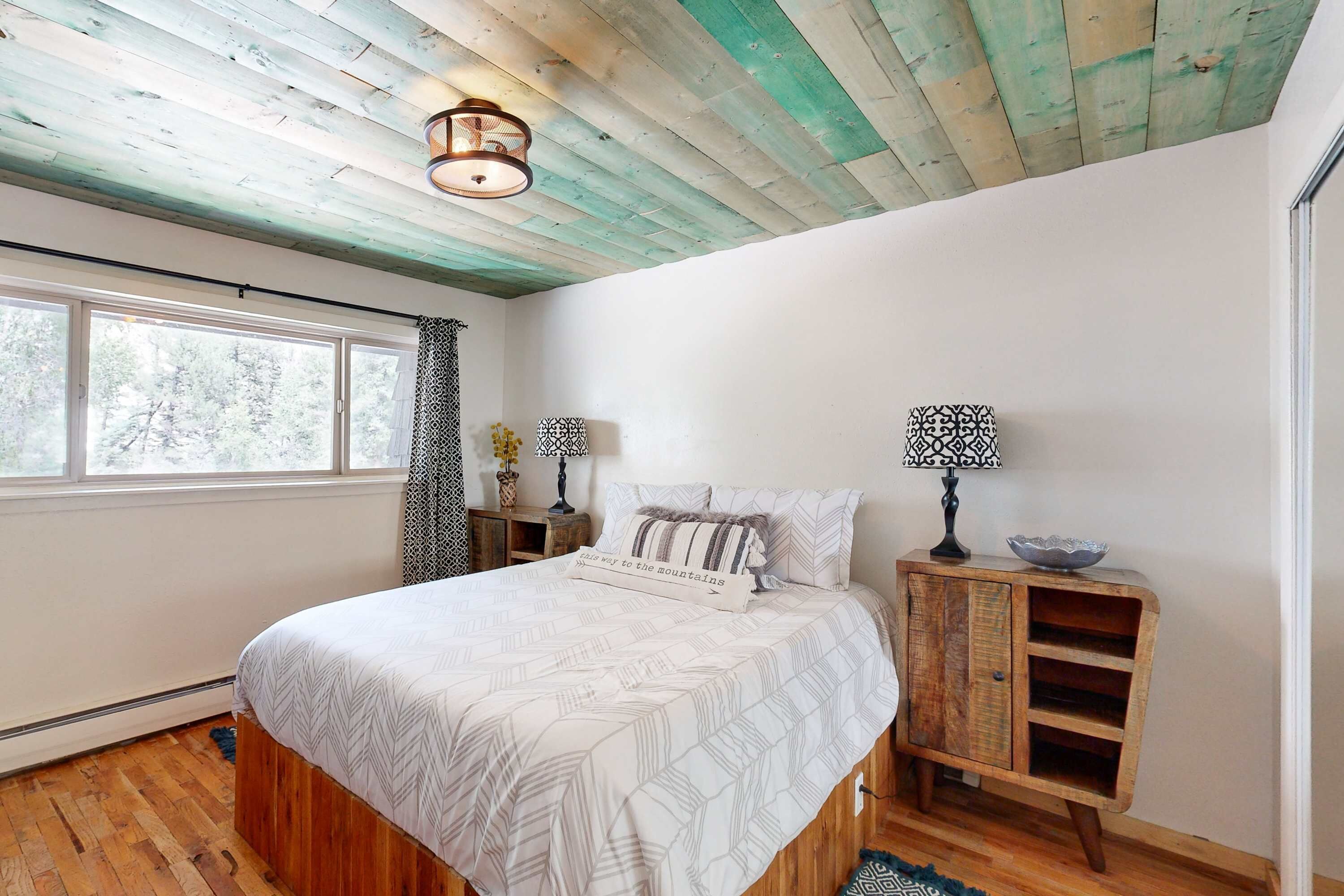 A bedroom with a bed, a nightstand, a lamp, a wooden cabinet, and a window with a view of trees.