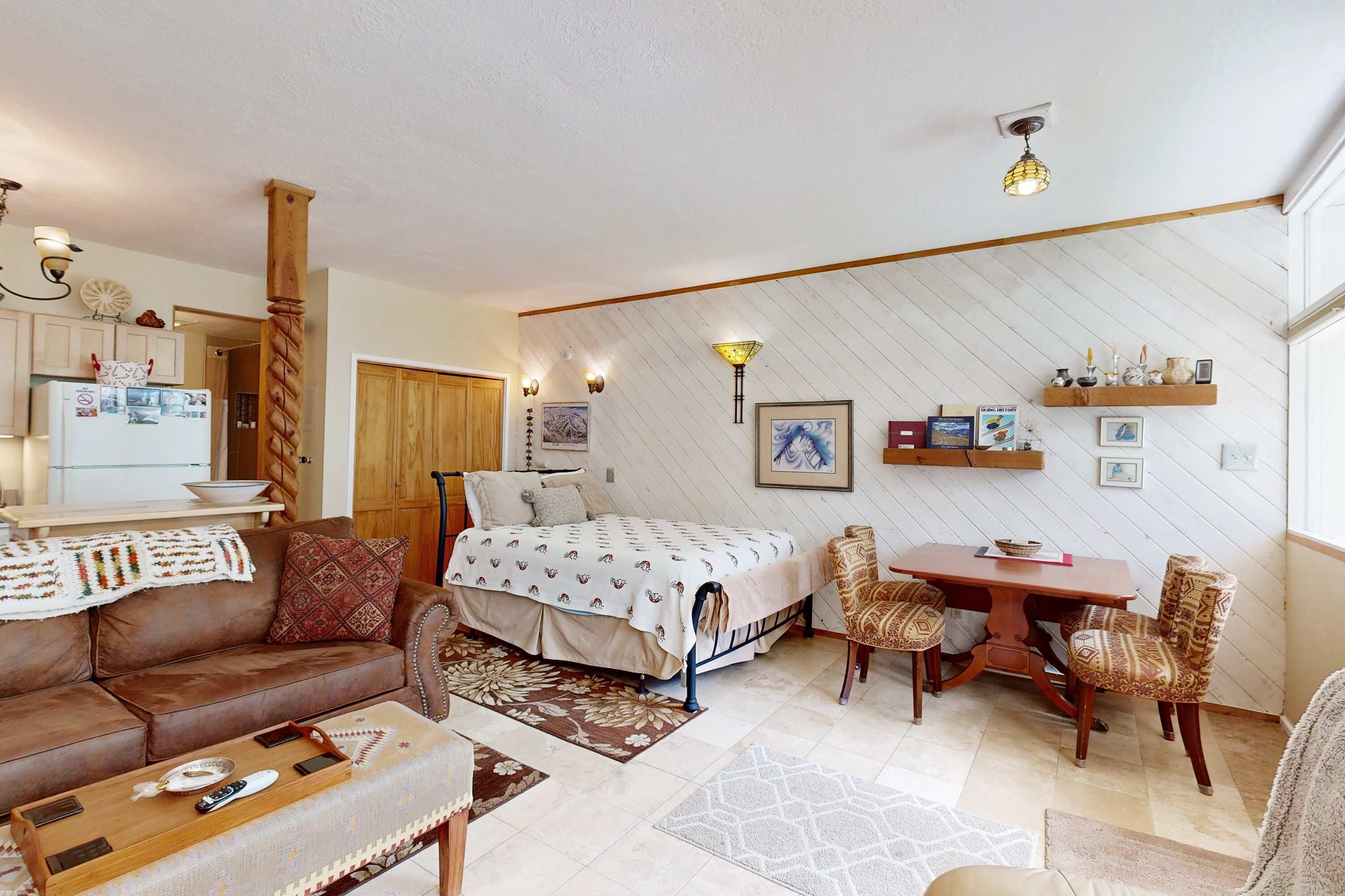 Una sala de estar acogedora con un sofá, una mesa de comedor con sillas, una cama con colcha estampada y una zona de cocina con refrigerador.