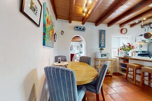 Un comedor con una mesa redonda de madera, sillas azules a rayas, una cocina al fondo y un reloj en la pared.