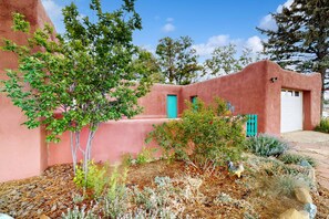 Un edificio de fachada rosa, con una puerta azul y un pequeño jardín con diversas plantas y una estatua de gallo.