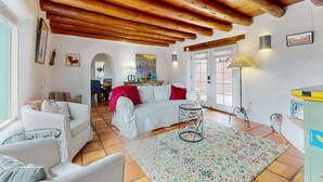 A living room with a white sofa, a glass coffee table, a patterned rug, wooden beams on the ceiling, and a dining area in the background. - The Birdhouse - Historic Adobe Architecture (Taos)