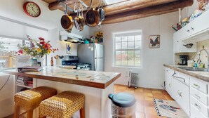A kitchen with a wooden ceiling, hanging pots, a clock, a refrigerator, and a window. - The Birdhouse - Historic Adobe Architecture (Taos)