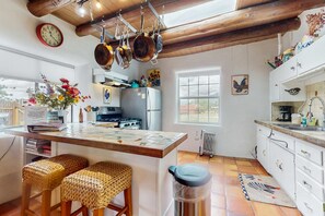 A kitchen with a wooden ceiling, hanging pots, a clock, a refrigerator, and a window. - The Birdhouse - Historic Adobe Architecture (Taos)