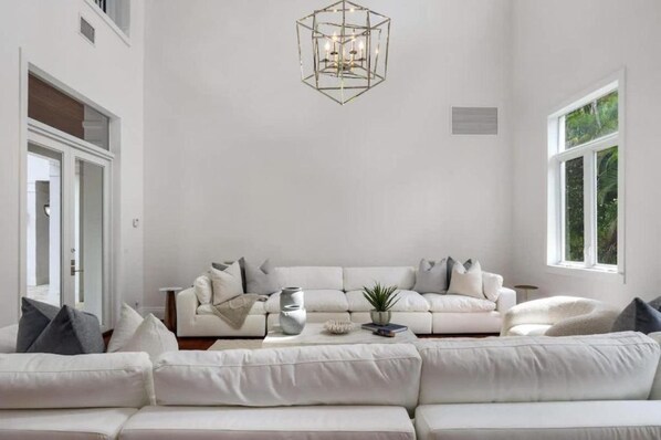 Un salon spacieux avec un ensemble de canapés blancs, un lustre aux motifs géométriques et une grande fenêtre laissant entrer la lumière naturelle.