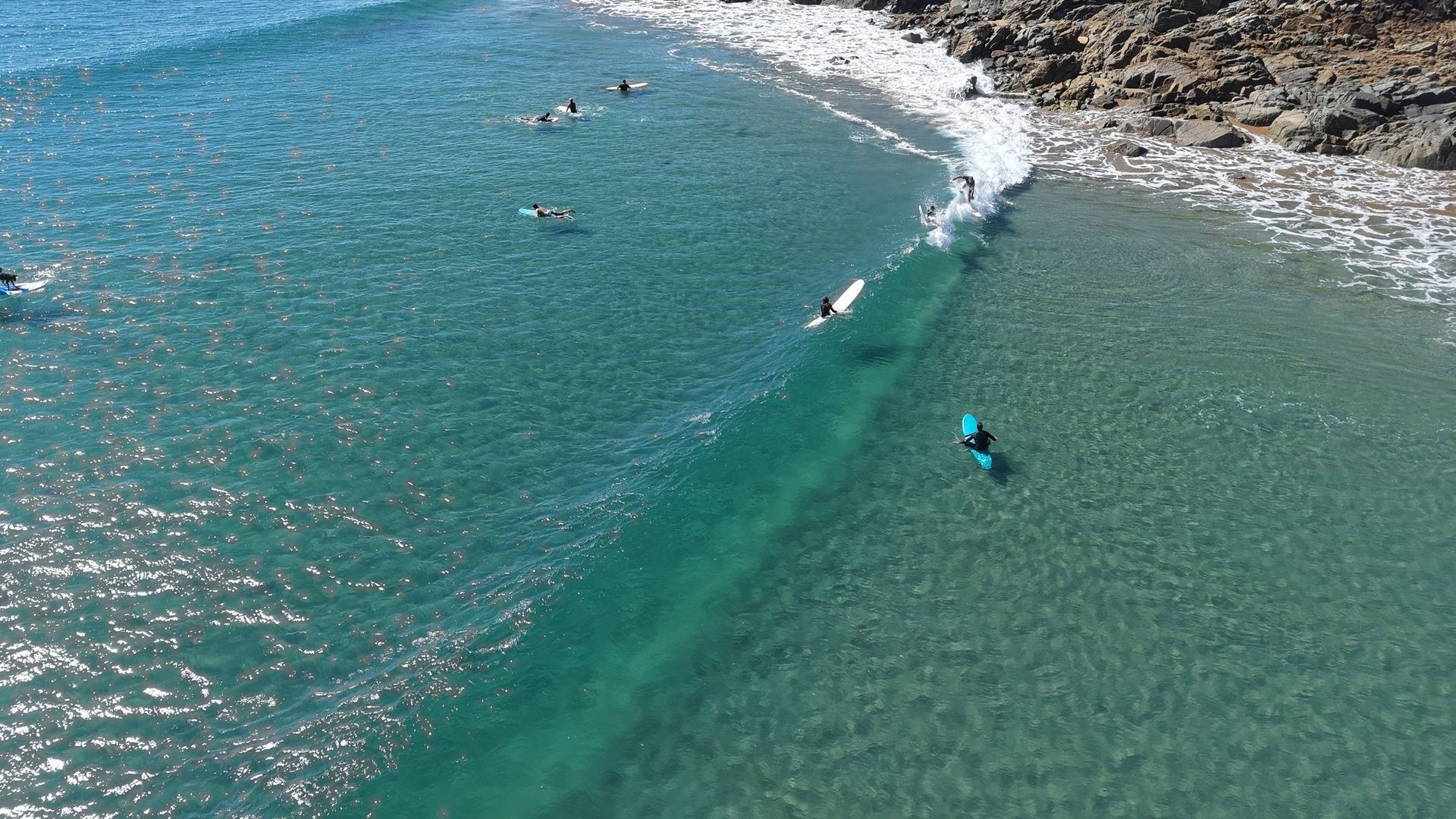Vista aérea de una playa con aguas turquesas cristalinas, costa rocosa y surfistas.