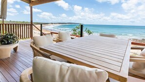 Een houten terras met een tafel en stoelen, met uitzicht op een strand, de oceaan en een verre kustlijn.