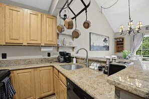 Une cuisine avec des meubles en bois, des plans de travail en granit, un évier en acier inoxydable, une machine à café et des casseroles suspendues.