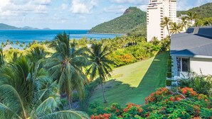 A hotel with a view of the ocean, a mountain, and a well-maintained lawn with palm trees and flowering plants.